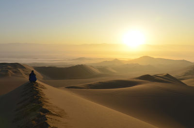 Rear view of man sitting on sand dune in desert against sky during sunrise