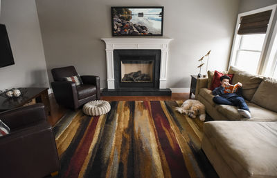 Teenage boy reading book on couch in living room with dog at his side.