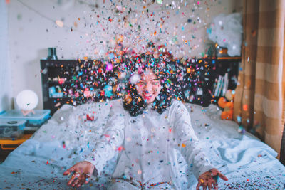 Happy young woman throwing confetti while sitting on bed at home