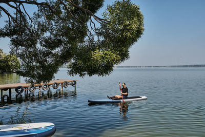 Asian older man sitting on sup board, rowing with oar on calm lake, floating to pier.