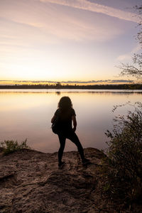 Rear view of woman standing by lake against sky during sunset