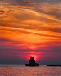 Silhouette buildings by sea against orange sky