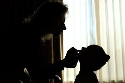 Side view of silhouette mother applying make-up on daughter face against curtain