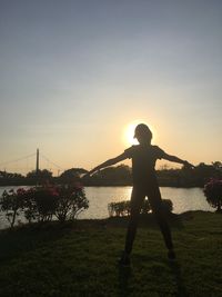 Silhouette boy standing at riverbank against sky during sunset