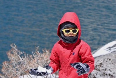 Boy in sunglasses and warm clothing holding snow while standing on lakeshore