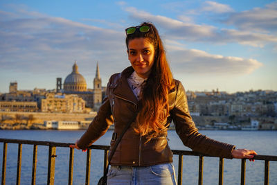 Portrait of young woman standing by railing against sky