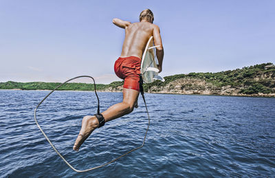 Shirtless man with surfboard jumping into sea against sky during sunny day