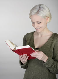 Smiling young woman reading book against wall