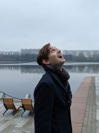 Side view of man with mouth open looking up while standing against river