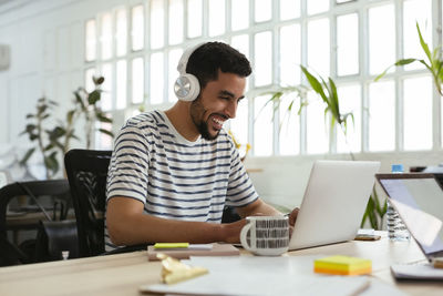 Laughing young man wearing headphones using laptop at desk in office