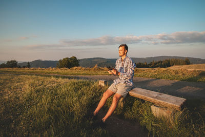 Photographer in a shirt sitting on top of a hill operates his drone to take pictures of  landscape