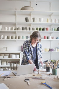 Mature female potter standing with laptop at table in workshop