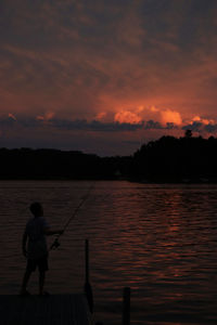 Silhouette boy fishing in lake against sky during sunset