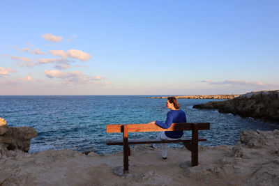 Man sitting on bench looking at sea shore against sky