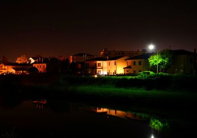 Illuminated buildings by lake against sky at night