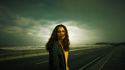 Portrait of young woman standing on road against cloudy sky