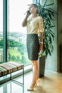 Full length of businesswoman talking on phone while standing by window