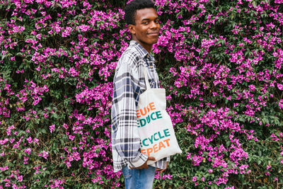 Side view of cheerful african american male standing with eco friendly shopping bag in blooming summer garden and looking at camera