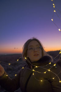 Young woman holding illuminated string light on field against sky during sunset