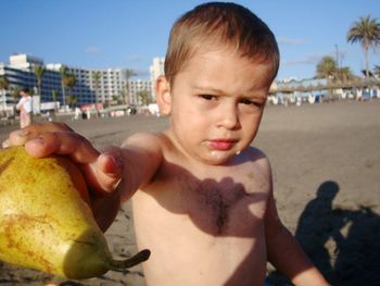 Portrait of shirtless boy holding pear at beach against sky