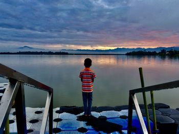 Rear view of boy standing on pier by sea during sunset