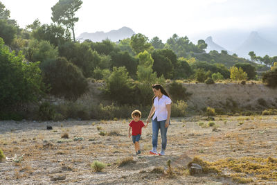 A woman and her young child holding hands while walking on a sunny nature trail with mountains