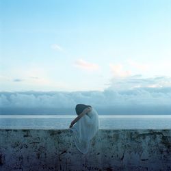 Woman sitting on surrounding wall against cloudy sky