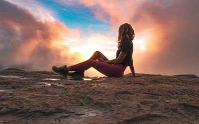 Woman sitting on rock against sky during sunset