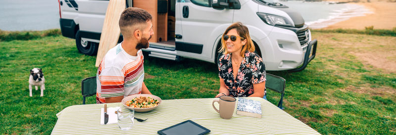 Couple sitting outdoors with camper van and beach on background