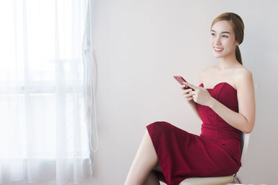 Beautiful young woman in red dress using smart phone while sitting against wall