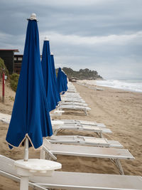 Deck chairs and closed parasols at beach against cloudy sky