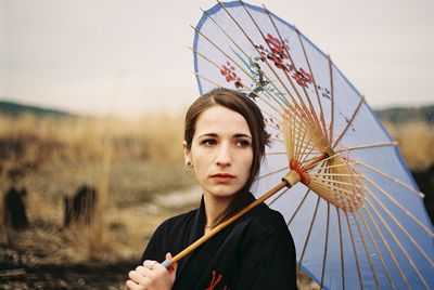 Thoughtful young woman looking away while holding paper umbrella against sky
