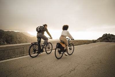 Male and female friends riding bicycles on road against sky