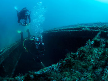 Professional divers swimming near old sunken ship covered with moss on bottom of deep sea with clear water