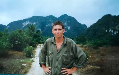 Portrait of mature man with arms akimbo standing on footpath against mountain