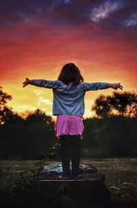 Rear view of girl with arms outstretched standing on field against sky during sunset