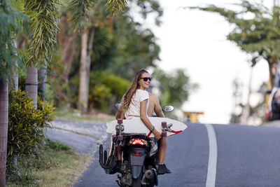 Rear view of friend riding motorcycle while woman holding skateboard