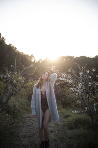 Full length of seductive young woman standing on land against sky at sunset