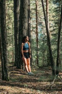 Tired young woman in sportswear leaned on a tree and resting in the forest after working out. 