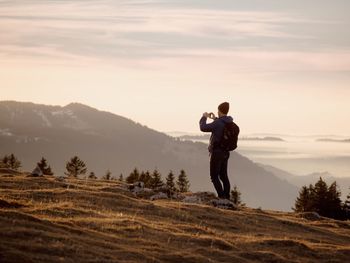 Backpacker photographing while standing on mountain against sky during sunset