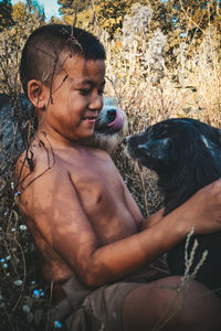 Shirtless boy playing with dogs while sitting against plants