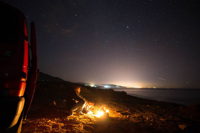 Man sitting by campfire on mountain against sky at night