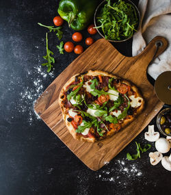 Top view of handmade pizza on wooden board with ingredients around it.