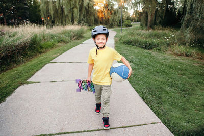 Portrait of boy holding skateboard walking on footpath at park