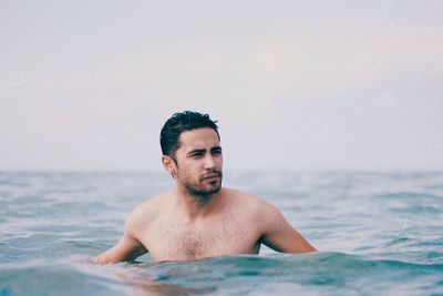 Shirtless young man looking away while swimming in sea against sky