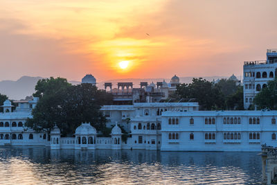 Buildings by river against sky during sunset