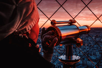 Woman holding coin operated binoculars by tour eiffel against sky during sunset