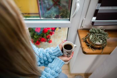 A woman sits at home holding a mug of hot coffee in her hands.