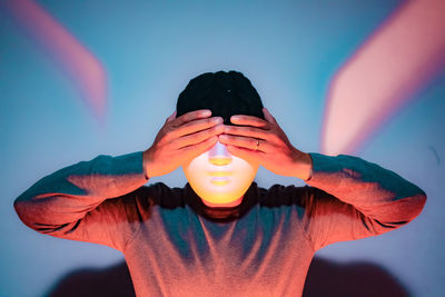 Close-up of man covering eyes while wearing mask against illuminated wall