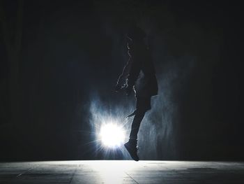 Full length side view of silhouette man jumping against illuminated spot light at night